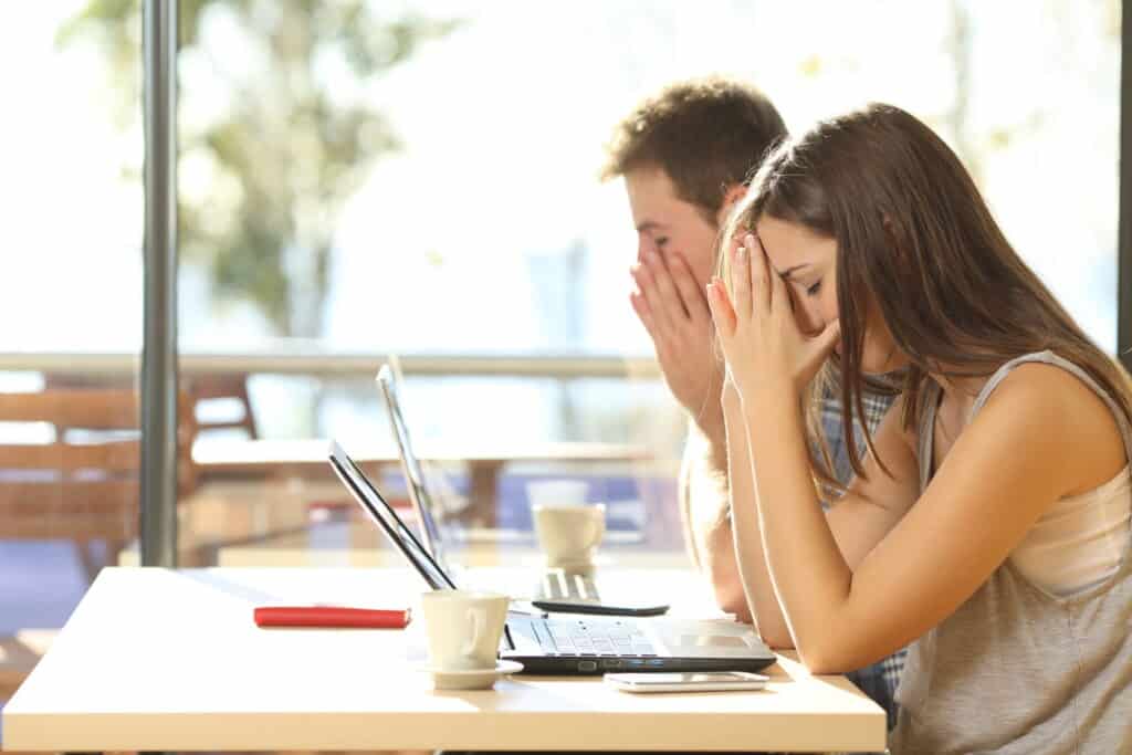 Ein Mann und eine Frau sitzen verzweifelt mit den Händen vorm Gesicht vor ihren Laptops.