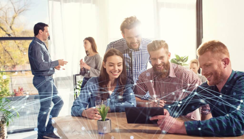 Colleagues are sitting at a large wooden table, looking at a tablet together and enjoying themselves.