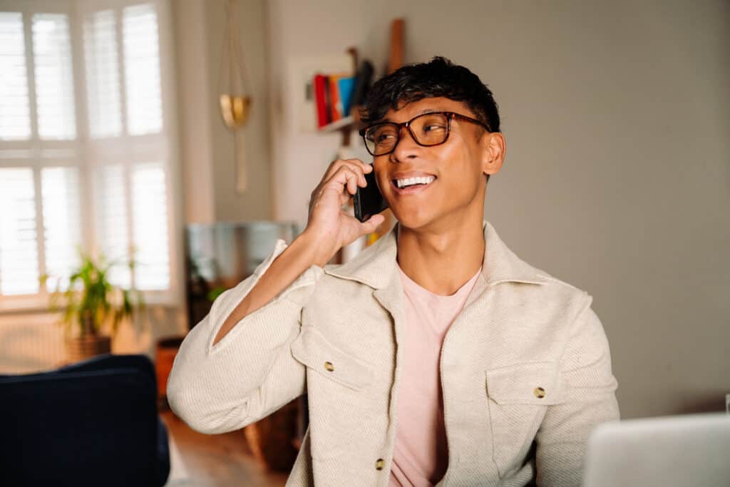 Ein junger Mann sitzt vor seinem Laptop und führt lächelnd ein Telefonat.