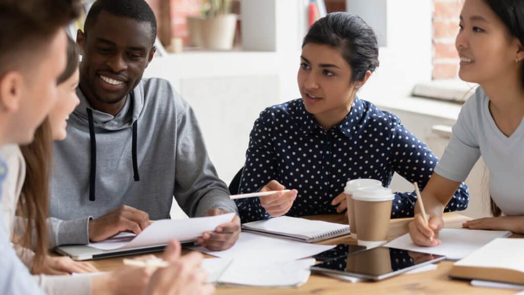 Fünf Arbeitskolleginnen und-Kollegen sitzen gemeinsam an einem Tisch mit Tablets, Notizblöcken und Kaffee, und beraten sich.